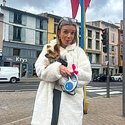 César participe au concours pour gagner de l'argent avec cette photo : woman, dog, urban, sidewalk, coat, playful, tongue_out, small_dog, city, buildings, flags, cloudy_sky, street, crosswalk, traffic_light, glasses, casual, leash, holding, outdoor