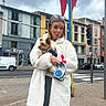 woman, dog, urban, sidewalk, coat, playful, tongue_out, small_dog, city, buildings, flags, cloudy_sky, street, crosswalk, traffic_light, glasses, casual, leash, holding, outdoor
