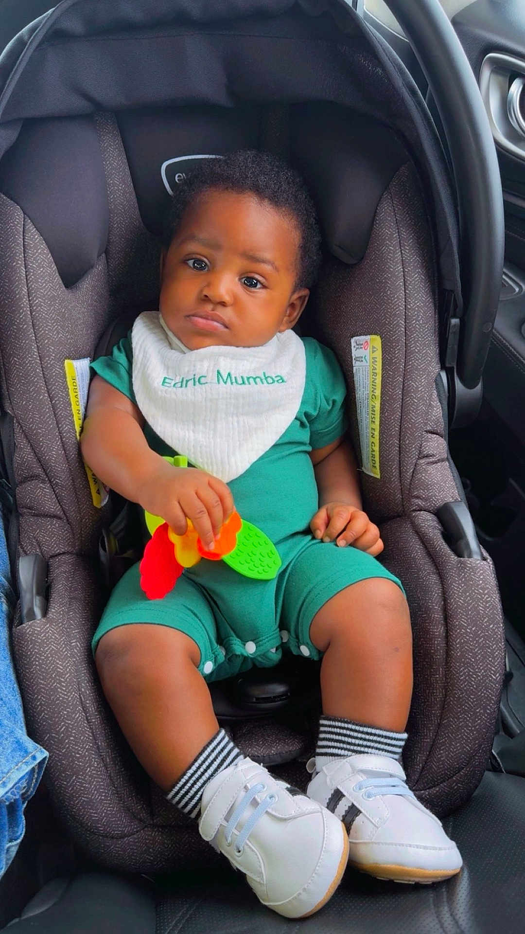 Edric joined the competition — help win amazing prizes! baby, child, car_seat, toy, bib, green_clothing, white_shoes, striped_socks, indoor, car_interior, serious_expression, infant, seatbelt, child_safety, person, cute, small_hands, black_hair, plush_seat, casual_wear