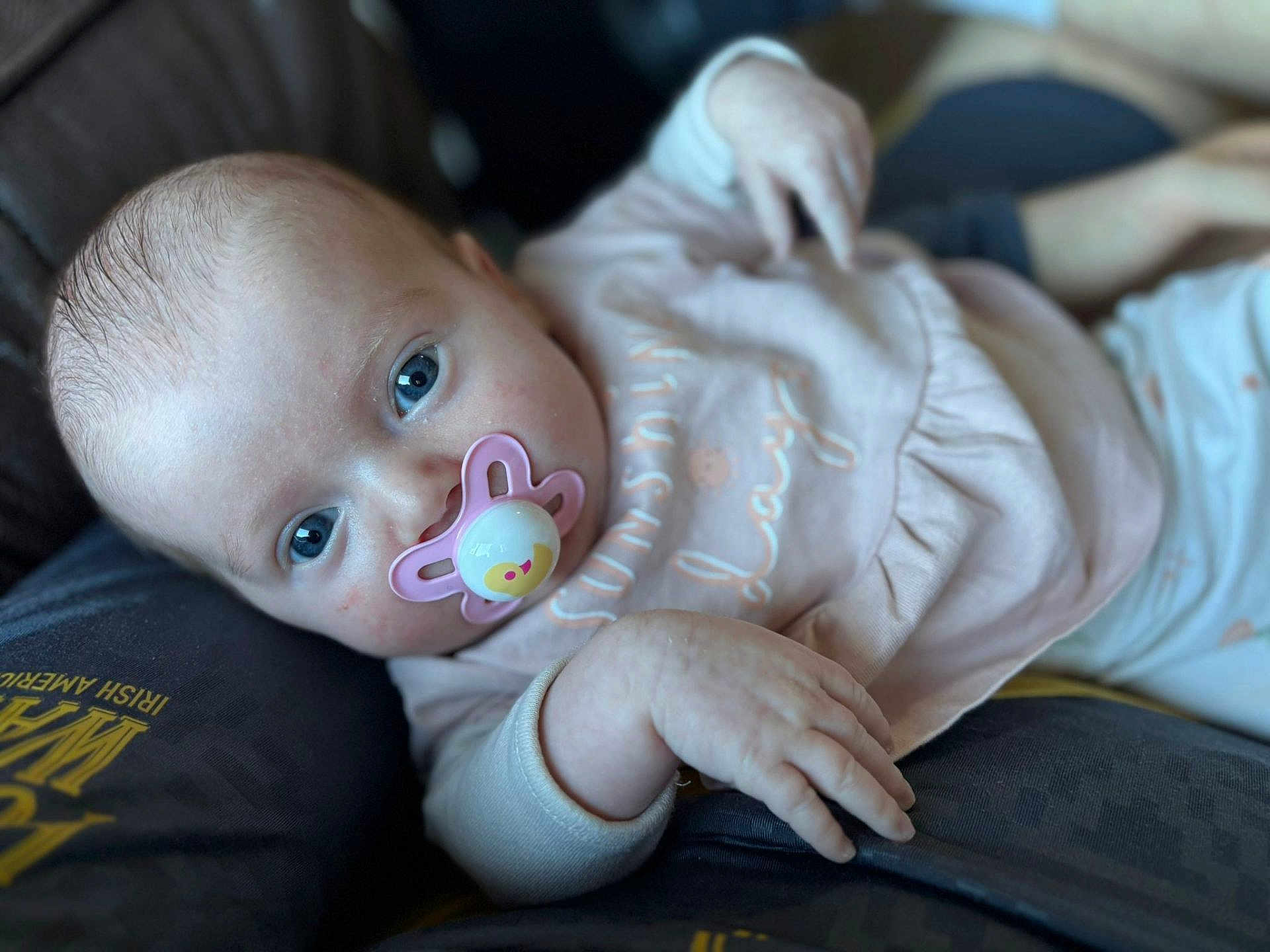 Daisy-Mae is registered to the contest to win money with this photo: baby, infant, pacifier, blue_eyes, pink_clothing, hand, face, close_up, lying_down, person, soft_focus, skin, cute, child, young, portrait, comfort, relaxing, indoor, human