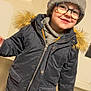 child, smiling, glasses, winter_jacket, knitted_hat, indoor, person, coat, fashion, cute, portrait, young_child, happy, casual, warm_clothing, fur_hood, standing, light_skin, headwear, zipped_jacket