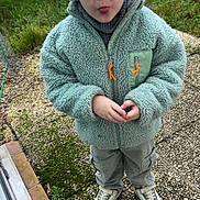 Marius participe au concours pour gagner de l'argent avec cette photo : child, boy, glasses, hat, fleece_jacket, green_jacket, outdoor, grass, moss, pathway, tongue_out, playful, sneaky, wall, concrete, sneakers, pants, standing, person, casual_clothing