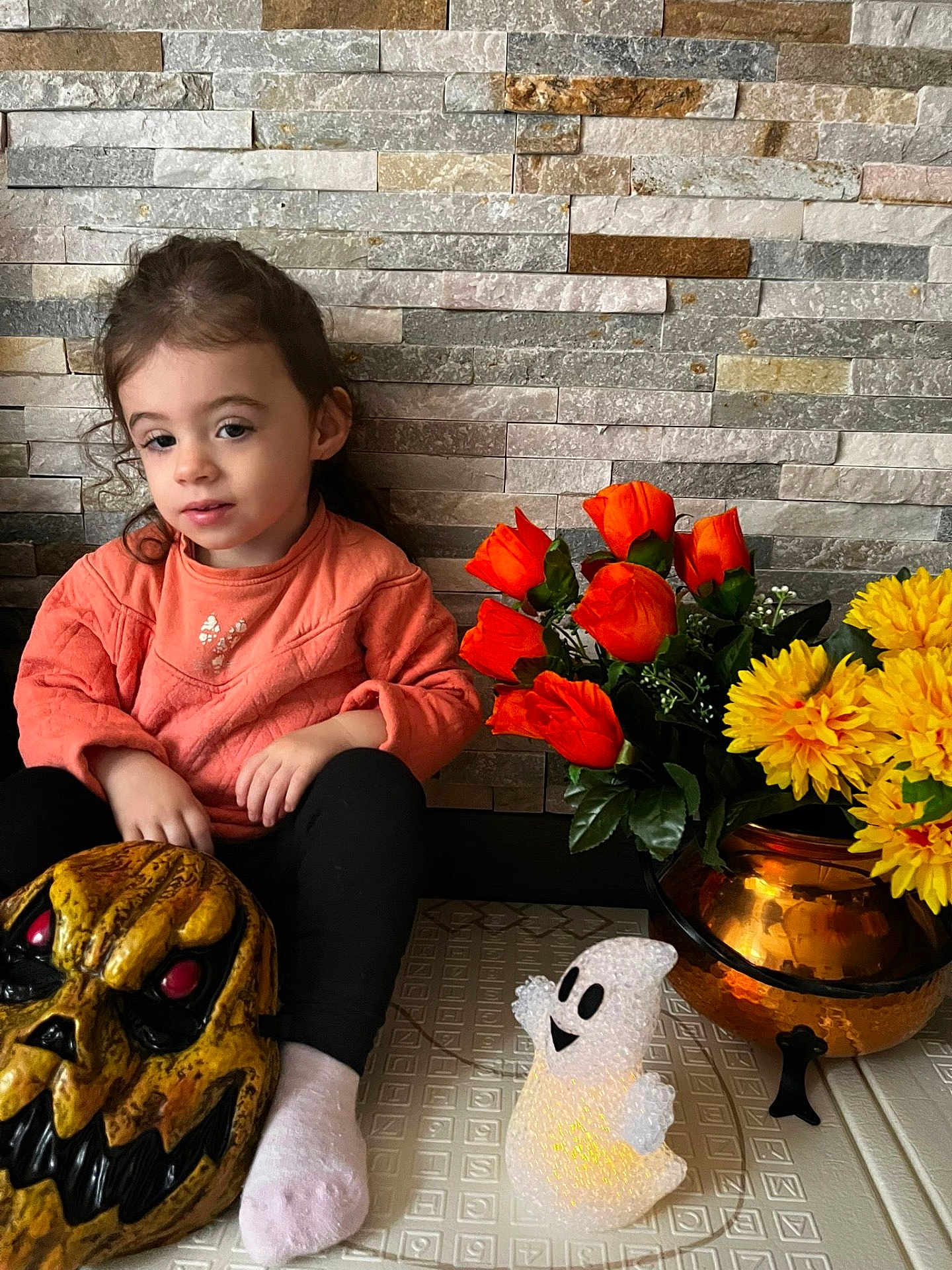 Laïa a rejoint le concours — aidez-le/la à gagner de superbes lots ! toddler, child, orange_sweater, black_pants, white_sock, halloween_mask, ghost_decoration, orange_flowers, yellow_flowers, flower_vase, copper_vase, stone_wall, indoor, sitting, toy, decoration, holiday, face, person, floor