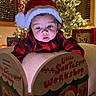 baby, child, santa_hat, book, reading, christmas_tree, holiday, plaid_clothing, indoors, festive, decorations, lights, face, portrait, cute, holiday_season, seasonal, warm_lighting, cozy, infant