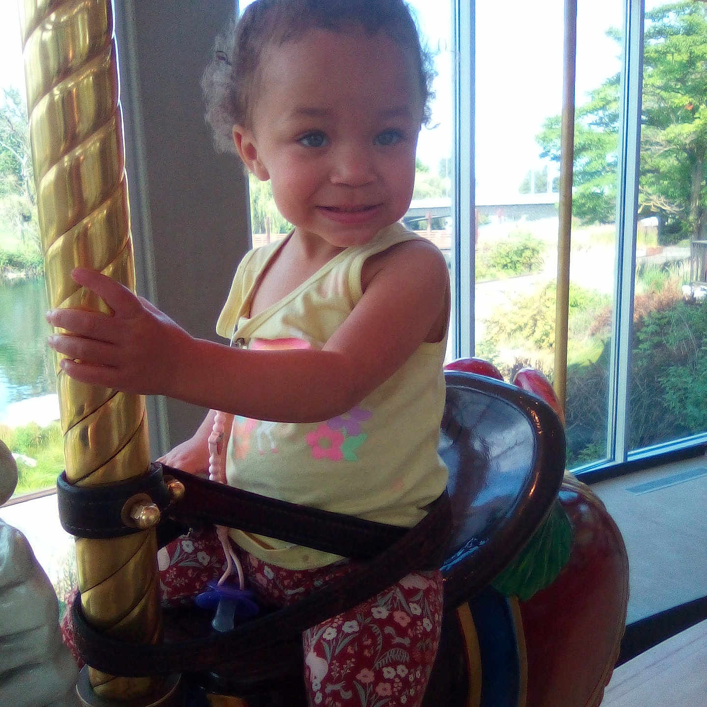 Jaicy is registered to the contest to win money with this photo: amusement_ride, carousel, casual_clothing, child, curly_hair, daylight, fun, golden_pole, happy, horse, indoor, patterned_pants, playful, portrait, seat, smiling, toddler, window, yellow_shirt, young_child