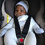 baby, car_seat, infant, child, white_clothing, hat, seat_belt, black_straps, cute, padded_seat, indoor, portrait, child_safety, young_child, infant_clothing, soft_lighting, baby_face, car_interior, baby_gloves, secure