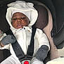 baby, child, car_seat, white_clothing, mittens, hood, face, infant, seatbelt, safety, cute, warm_clothing, portrait, young_child, indoors, black_skin, baby_outfit, baby_accessories, cozy, baby_hat