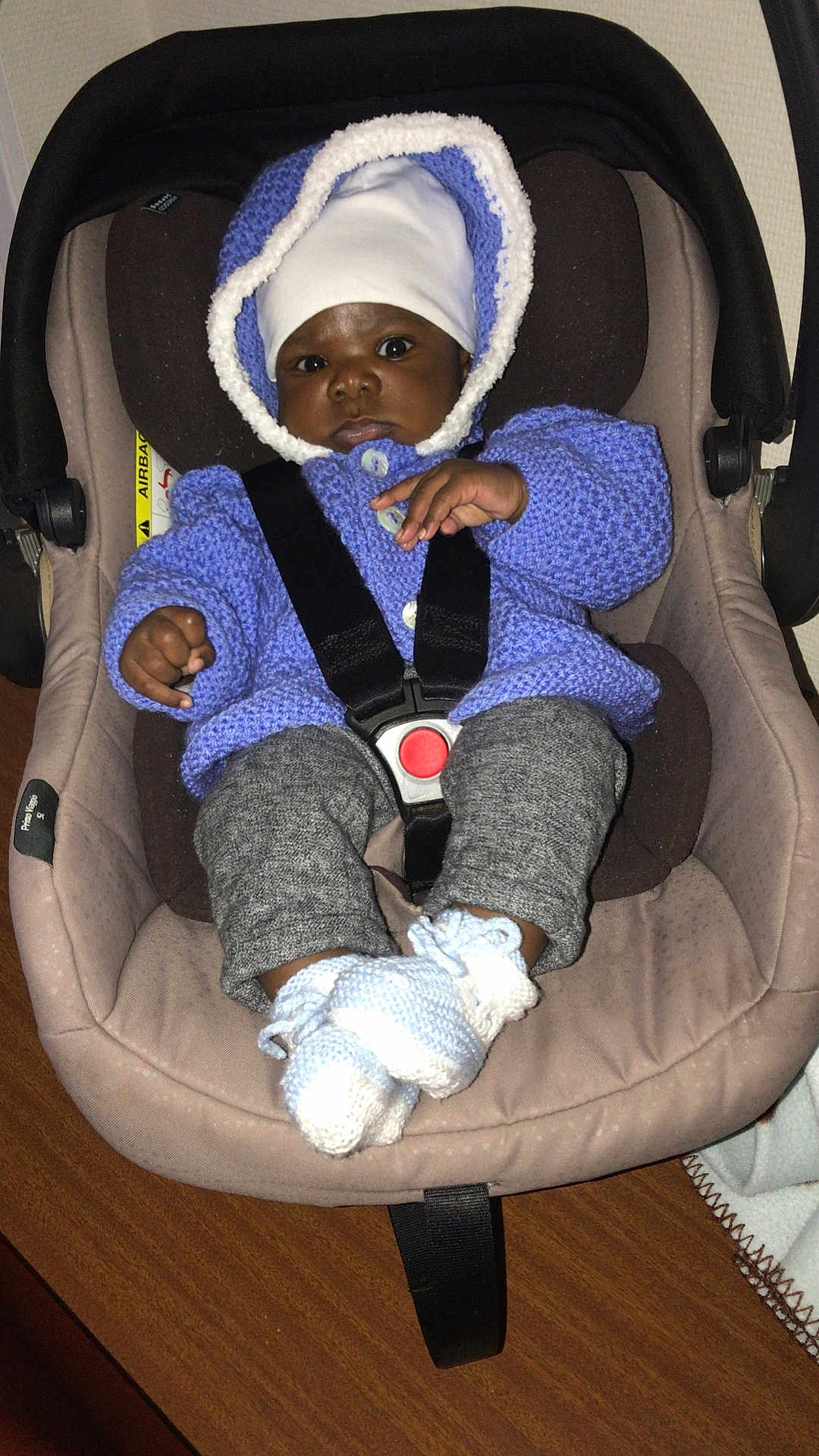 Nathael Angel a rejoint le concours — aidez-le/la à gagner de superbes lots ! baby, car_seat, knitted_sweater, hat, booties, infant, child, seatbelt, indoor, wooden_table, curious, black_skin, face, clothing, portrait, cute, warm_clothes, hands, legs, comfort