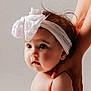 baby, infant, headband, bow, big_eyes, portrait, studio_lighting, cute, chubby_cheeks, hand, supporting_hand, skin, face, hair, looking_away, expression, soft_background, child, newborn, close_up