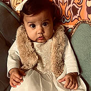 Charlotte a rejoint le concours — aidez-le/la à gagner de superbes lots ! baby, child, dress, fur_vest, bow, cute, wide_eyes, patterned_background, indoor, seated, portrait, furniture, hand, curious, colorful, person, infant, decor, fabric, expression
