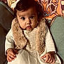 baby, child, dress, fur_vest, bow, cute, wide_eyes, patterned_background, indoor, seated, portrait, furniture, hand, curious, colorful, person, infant, decor, fabric, expression