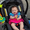 baby, infant, car_seat, stroller, toy, lemon, blue_clothing, pink_skirt, cute, outdoor, child, pavement, footwear_free, sitting, face, expression, colorful, plastic_toy, happy, young