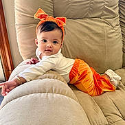 Taelynn joined the competition — help win amazing prizes! baby, child, couch, orange_pants, headband, bow, white_top, indoors, plush, relaxing, cute, smiling, infant, soft_furniture, cozy, portrait, person, young_child, comfortable, home