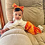 baby, child, couch, orange_pants, headband, bow, white_top, indoors, plush, relaxing, cute, smiling, infant, soft_furniture, cozy, portrait, person, young_child, comfortable, home