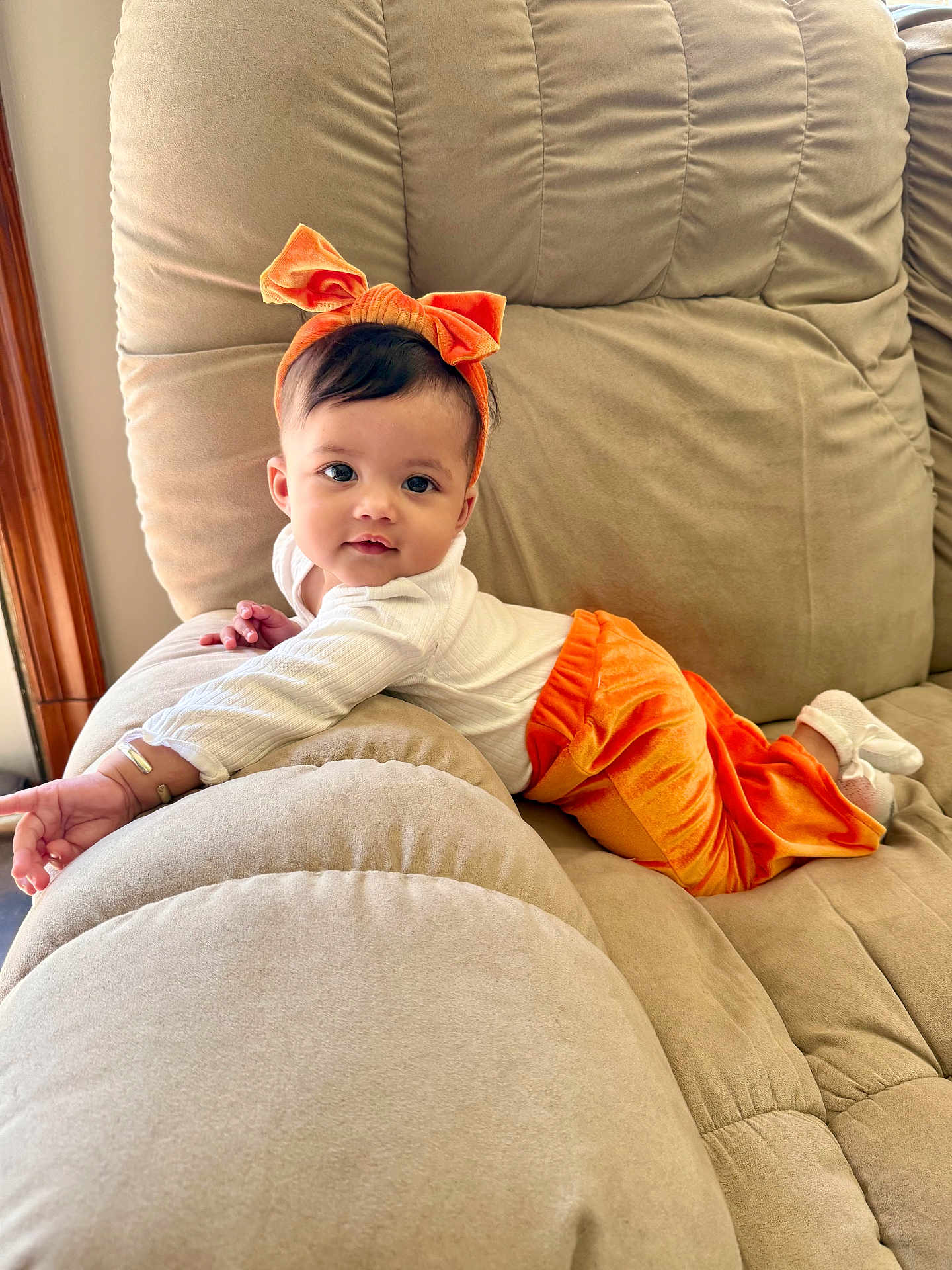 Taelynn joined the competition — help win amazing prizes! baby, child, couch, orange_pants, headband, bow, white_top, indoors, plush, relaxing, cute, smiling, infant, soft_furniture, cozy, portrait, person, young_child, comfortable, home