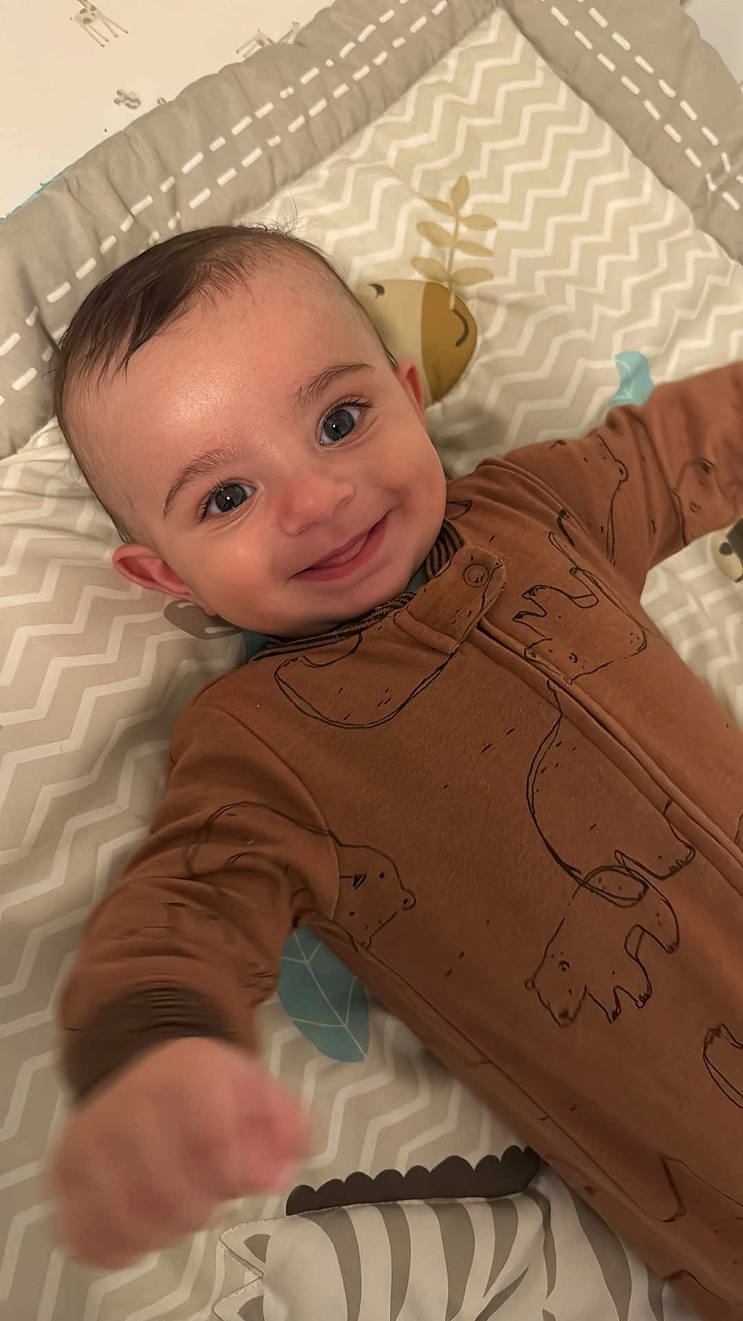 Christiano is registered to the contest to win money with this photo: baby, smiling, onesie, brown_clothing, playmat, patterned, face, infant, happy, child, cute, reaching, lying_down, indoors, person, portrait, head, closeup, baby_gesture, soft_lighting