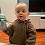 toddler, child, baby, hoodie, sippy_cup, indoor, living_room, television, wooden_floor, furniture, table, person, young_child, cozy, casual_clothing, curious, expression, holding, standing, home