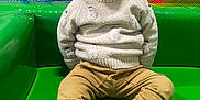 Valentin a rejoint le concours — aidez-le/la à gagner de superbes lots ! toddler, child, sitting, green_surface, sweater, pants, socks, indoor, play_area, netting, colorful, face, person, young_child, casual_clothing, footwear, baby, portrait, seated, expression