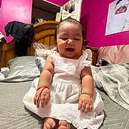 Marilu joined the competition — help win amazing prizes! baby, child, white_dress, bow, bed, pillow, blanket, wooden_headboard, pink_wall, smiling, happy, indoor, portrait, person, footwear, bracelet, red_string, photograph, cozy, room
