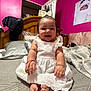 baby, infant, smiling, dress, white_dress, bow, bed, blanket, pink_wall, portrait, happy, cute, indoors, child, person, feet, bracelet, wooden_headboard, fabric, room