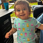 Woody is registered to the contest to win money with this photo: toddler, child, smile, curly_hair, colorful_clothing, patterned_outfit, bunny_pattern, chick_pattern, outdoor, table, cup, vehicle, grass, person, jeans, concrete_floor, happy, portrait, daylight, playful