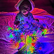Tariq is registered to the contest to win money with this photo: blanket, celebration, child, christmas_lights, colorful_lights, curious, festive, footwear, glowing, hat, holiday, indoors, light_effect, night, pajamas, person, playing, shoes, sitting, toddler
