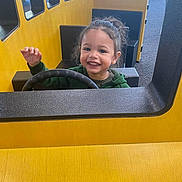 Aiden is registered to the contest to win money with this photo: child, toddler, smiling, playground, steering_wheel, yellow, green_clothing, indoor, play_structure, happy, curly_hair, seat, window, hand_raised, face, person, fun, play, young, toy