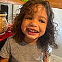 child, smiling, curly_hair, teeth, gray_shirt, kitchen, cabinet, countertop, tile_floor, happy, portrait, indoor, person, cute, young, playful, joyful, toddler, casual