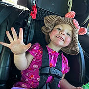 River is registered to the contest to win money with this photo: child, toddler, car_seat, hat, knitted_hat, pink_dress, cherry_pattern, smiling, happy, waving_hand, seatbelt, car_interior, toy, playtime, cute, portrait, face, skin, cheeks, hair