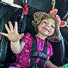 child, toddler, car_seat, hat, knitted_hat, pink_dress, cherry_pattern, smiling, happy, waving_hand, seatbelt, car_interior, toy, playtime, cute, portrait, face, skin, cheeks, hair