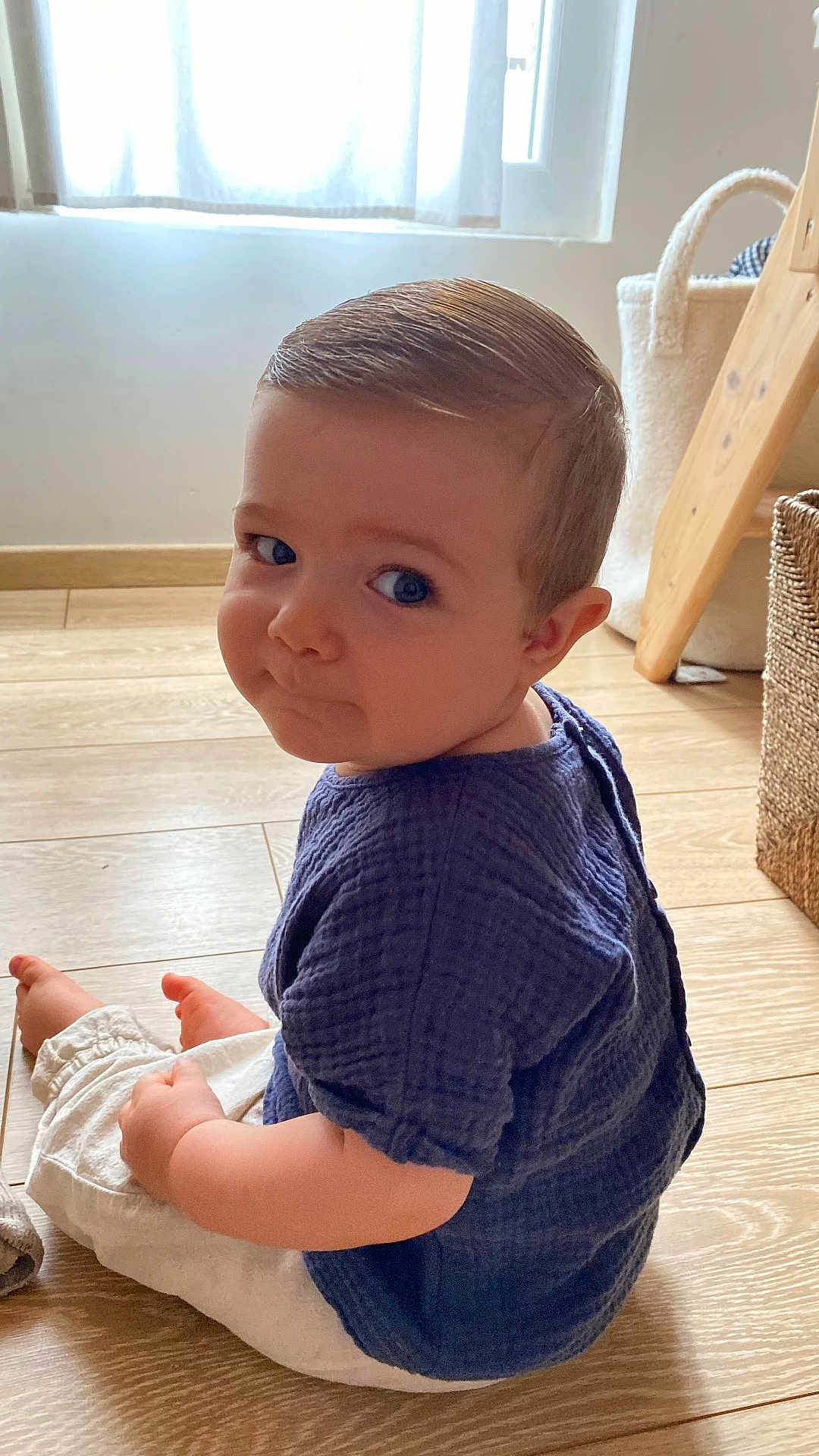 Léonard participe au concours pour gagner de l'argent avec cette photo : toddler, child, sitting, wooden_floor, blue_shirt, white_pants, indoor, natural_light, basket, curtain, window, flooring, looking_back, short_hair, cute, baby, person, home, expression, casual_clothing