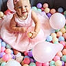 baby, child, pink_dress, bow, pearls, ball_pit, colorful_balls, balloons, smiling, happy, indoors, playful, cute, sitting, portrait, soft_lighting, pastel_colors, footwear, celebration, fun