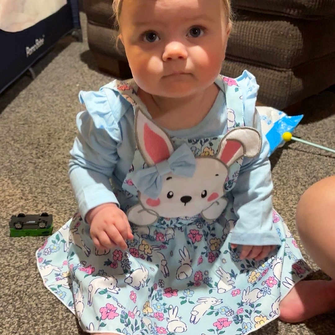 Maeve joined the competition — help win amazing prizes! baby, blue_dress, bunny_print, carpet, child, clothing, cute, expression, face, foot, furniture, hand, headband, home, indoor, person, sitting, sofa, toddler, toy