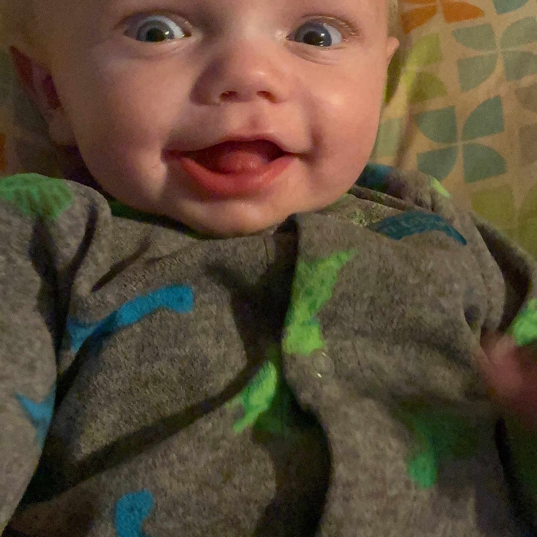 Elias Martin is registered to the contest to win money with this photo: baby, child, closeup, clothing, cozy, cute, dinosaur_print, expression, eyes, face, happy, indoor, infant, onesie, pattern, person, pillow, portrait, sleepwear, smiling