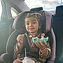 child, car_seat, donut, blue_frosting, drink_cup, car_interior, seatbelt, smiling, brown_outfit, bracelet, window, daylight, happy, food, snack, young_child, hair_tied, casual_clothing, sitting, cute
