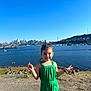 child, girl, green_clothing, peace_sign, smile, outdoor, water, skyline, city, daylight, grass, people, hill, recreation, summer, casual, portrait, blue_sky, nature, happy