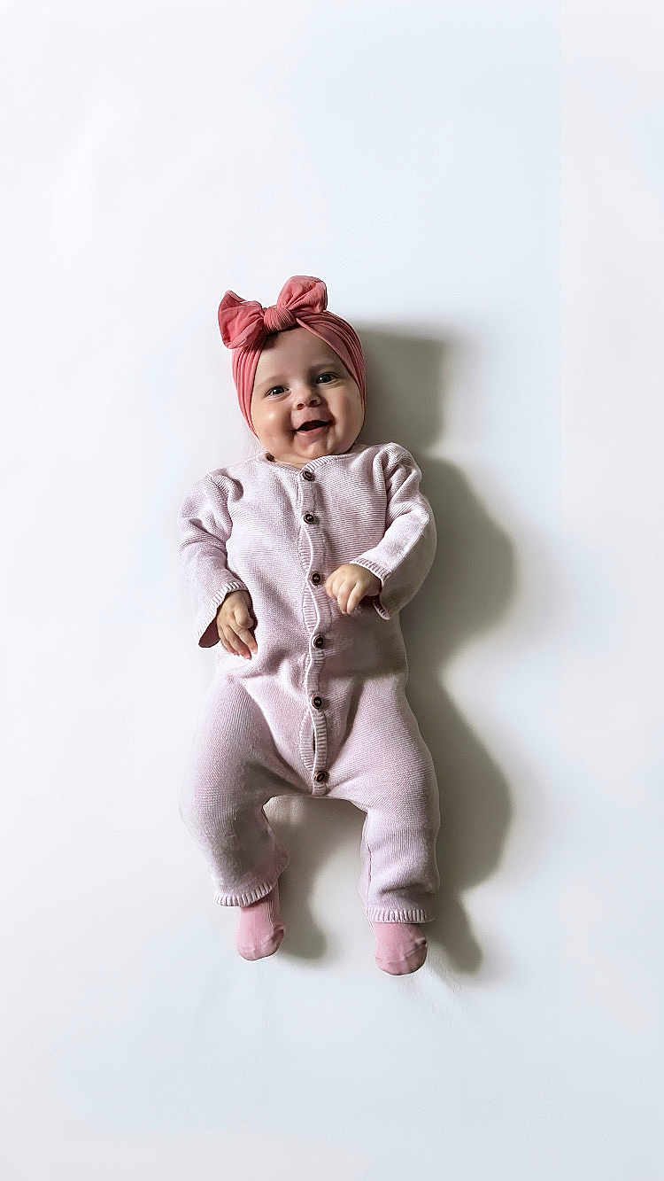Mia a rejoint le concours — aidez-le/la à gagner de superbes lots ! baby, infant, smiling, pink_onesie, headband, bow, lying_down, shadow, white_background, cute, child, portrait, happy, soft_texture, buttoned_clothing, newborn, small_hands, tiny_feet, studio_photo, adorable