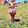 child, toddler, toy, stuffed_animal, purple, flowers, bluebonnets, field, grass, outdoor, sunny, smiling, red_barn, people, sky, nature, summer, casual_clothing, pink_shoes, happy