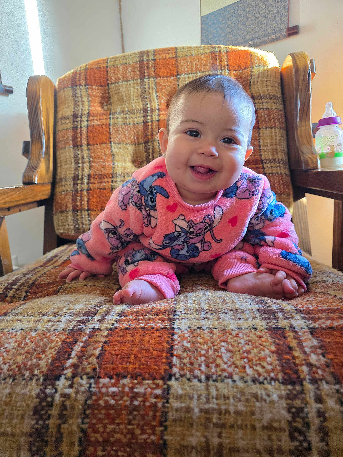 Kaori Delgado is registered to the contest to win money with this photo: baby, child, smiling, crawling, pink_clothing, cartoon_print, armchair, plaid_pattern, orange, brown, wooden_armrest, indoor, cozy, sunlight, happy, face, person, cute, foot, furniture
