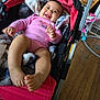 baby, smiling, pink_onesie, stroller, blanket, indoor, wooden_floor, happy, child, infant, person, footwear_absent, hand, face, head, furniture, toy, home, cozy, relaxing