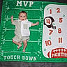 baby, blanket, celebration, child, clothing, cute, football, green, happy, helmet, indoor, infant, laying_down, milestone_blanket, months, numbers, person, portrait, smile, white
