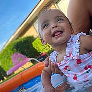 Brexley is registered to the contest to win money with this photo: toddler, child, swimsuit, pool, water, splashing, summer, outdoor, sunny, happy, smiling, baby, inflatable_pool, person, adult_hand, greenery, blue_sky, play, fun, warm_weather