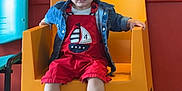 Diego Pieddefer a rejoint le concours — aidez-le/la à gagner de superbes lots ! child, toddler, orange_chair, red_outfit, denim_jacket, indoor, colorful_chairs, red_wall, floor_tiles, wooden_table, sitting, curious, casual_clothing, footwear, young_child, portrait, playful, bright_colors, furniture, person