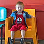 child, toddler, orange_chair, red_outfit, denim_jacket, indoor, colorful_chairs, red_wall, floor_tiles, wooden_table, sitting, curious, casual_clothing, footwear, young_child, portrait, playful, bright_colors, furniture, person
