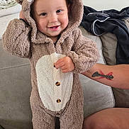 Zayden joined the competition — help win amazing prizes! baby, child, toddler, bear_costume, onesie, hood, fuzzy_clothing, smiling, standing, portrait, indoor, couch, sofa, adult_arm, tattoo, home, living_room, black_socks, buttoned_outfit, cute