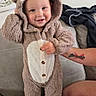 baby, child, toddler, bear_costume, onesie, hood, fuzzy_clothing, smiling, standing, portrait, indoor, couch, sofa, adult_arm, tattoo, home, living_room, black_socks, buttoned_outfit, cute