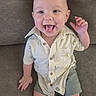 baby, child, infant, smiling, happy, sitting, couch, portrait, indoor, face, hand, waving, open_mouth, short_hair, shirt, patterned_clothing, shorts, barefoot, cushion, candid