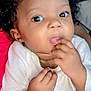 Samantha is registered to the contest to win money with this photo: baby, child, close_up, curly_hair, cute, expression, face, fingers, hand, indoor, infant, looking_at_camera, person, portrait, red_background, skin, sucking_fingers, toddler, white_onesie, young_child