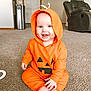 baby, infant, orange_hoodie, pumpkin_costume, smiling, sitting, carpet, living_room, furniture, cozy, hood, cute, child, indoors, happy, face, feet, home, playful, casual