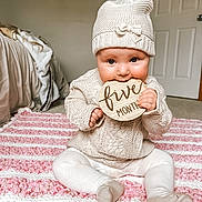 Brae is registered to the contest to win money with this photo: baby, blanket, carpet, child, clothing, cozy, cute, face, five_months, hands, indoor, infant, knit_hat, legs, pink, playful, pom_pom, sitting, white, wooden_sign